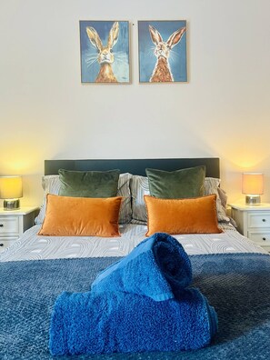 Een bed met beddengoed met een patroon, twee kussens, een blauwe deken en twee aan de muur hangende afbeeldingen van konijnen.