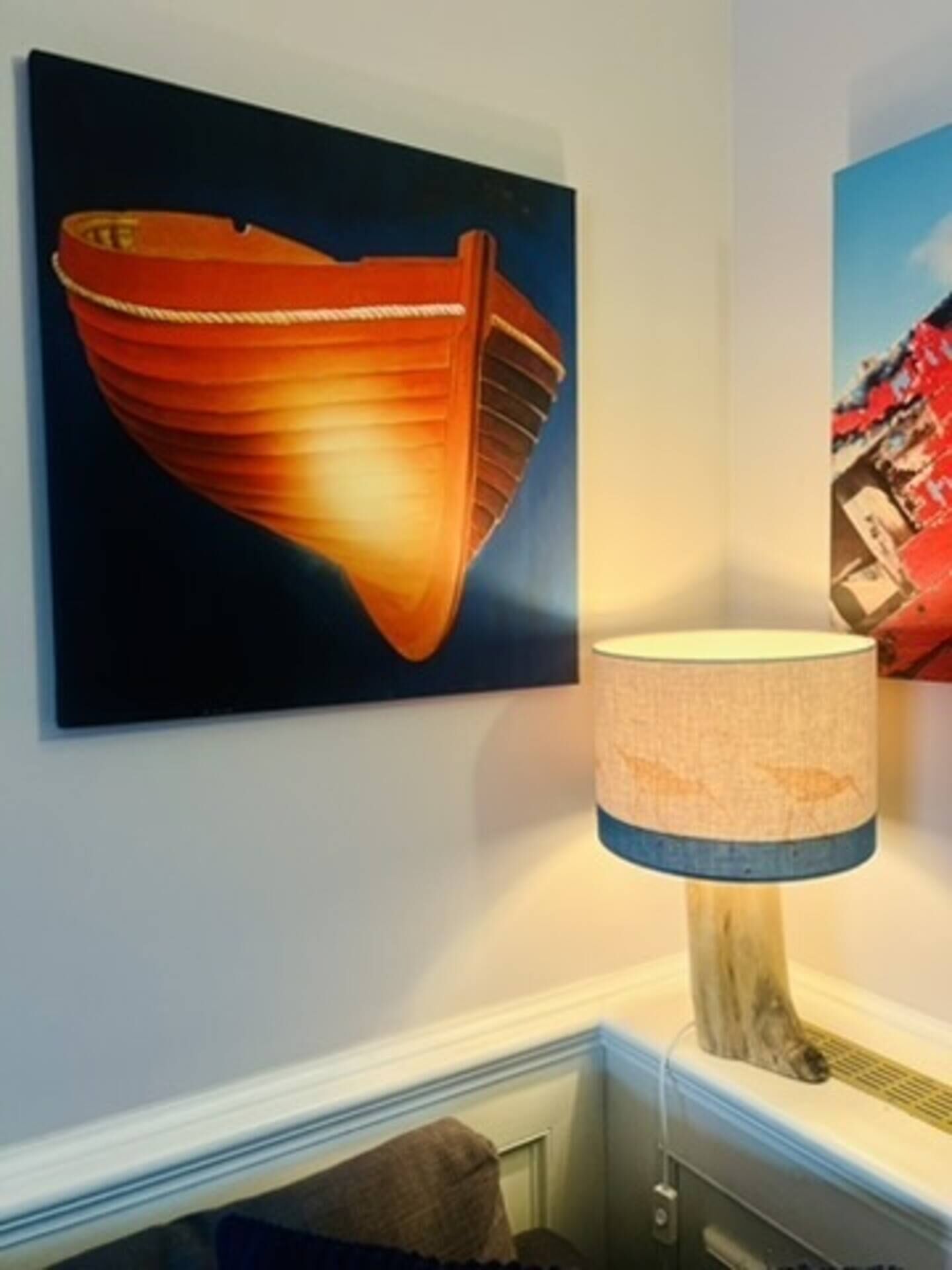 Une pièce dans laquelle on peut voir un tableau encadré représentant un bateau, une lampe avec un pied bleu et une vue partielle d’une fenêtre avec un rideau.