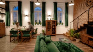 Een moderne woonkamer met een groene bank, een houten eettafel en stoelen. Er is een trap met een bakstenen muur en een televisie op een houten dressoir.