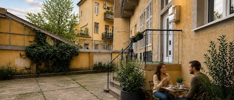 An outdoor patio area with a table and chairs, surrounded by potted plants and a building with balconies.