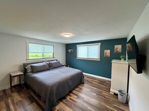 Een slaapkamer met een bed, twee ramen, een nachtkastje, een dressoir en een plafondlamp.