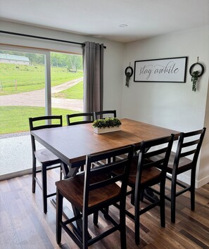 Een eetruimte met een houten tafel en stoelen, uitzicht op een groen landschap door een raam, en een lijst met een bord op de muur.