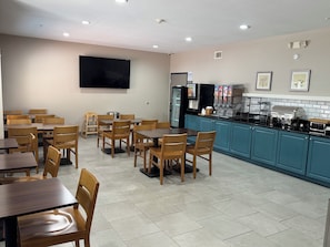 Ein Restaurant mit Holztischen und -stühlen, einem Küchenbereich mit einer Kaffeemaschine und einem an der Wand befestigten Flachbildfernseher.