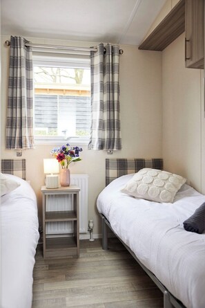 Une chambre avec deux lits, une table de chevet avec une lampe et un vase, des rideaux à carreaux et une fenêtre donnant sur des arbres.