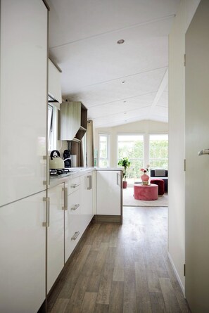 A modern kitchen with white cabinetry, a gas stove, and a view to a living area with a red ottoman and a plant. - 4P Modern Chalet with Private Sauna - Fenced Garden in a Forest Area - BBQ (Putten)