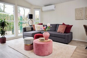 Un salon avec un canapé gris, des coussins rouges et blancs, un pouf rose, un vase rempli de fleurs et une grande fenêtre donnant sur un paysage verdoyant.