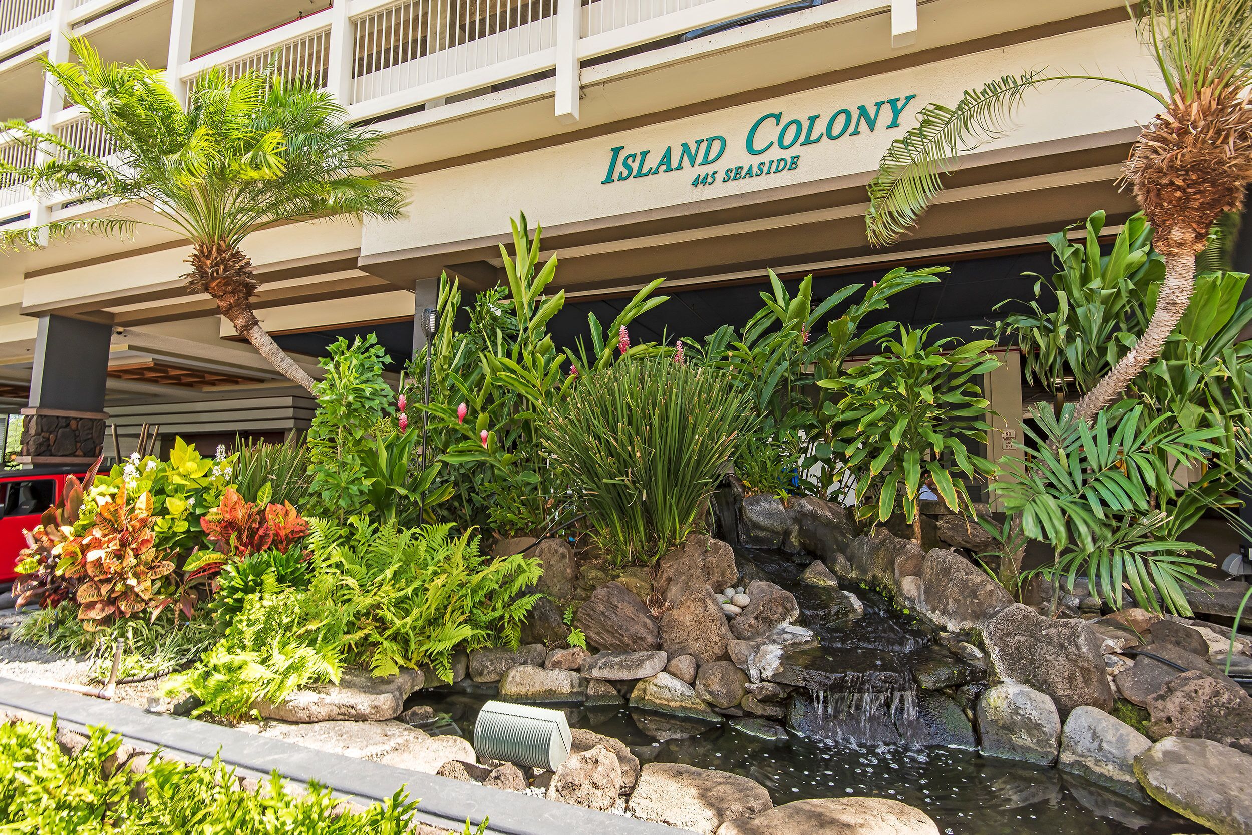 A hotel named 'Island Colony' with a landscaped garden featuring a small waterfall and various tropical plants.