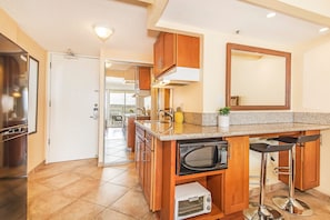 A kitchen with wooden cabinets, a microwave, a toaster, and a bar stool. - Ocean-diamond Head Views From Island Colony Condo Short Walk to Waikiki Beach (Honolulu)