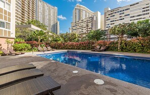 A swimming pool with lounge chairs, surrounded by greenery and tall buildings. - The Cove Waikiki Condo W/full Kitchen by the Beach and Free Parking/wifi (Honolulu)