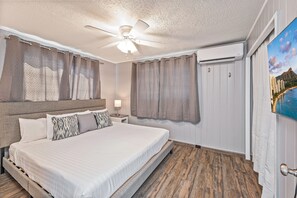 Una habitación de hotel con cama, cortinas, ventilador de techo, equipo de aire acondicionado y un cuadro de un paisaje.