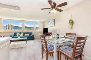 A modern living room with a dining area, large windows with a city view, a ceiling fan, and a flat-screen TV. - Mountain & Golf Course Views Family Island Colony Condo & Lots of Amenities (Honolulu)