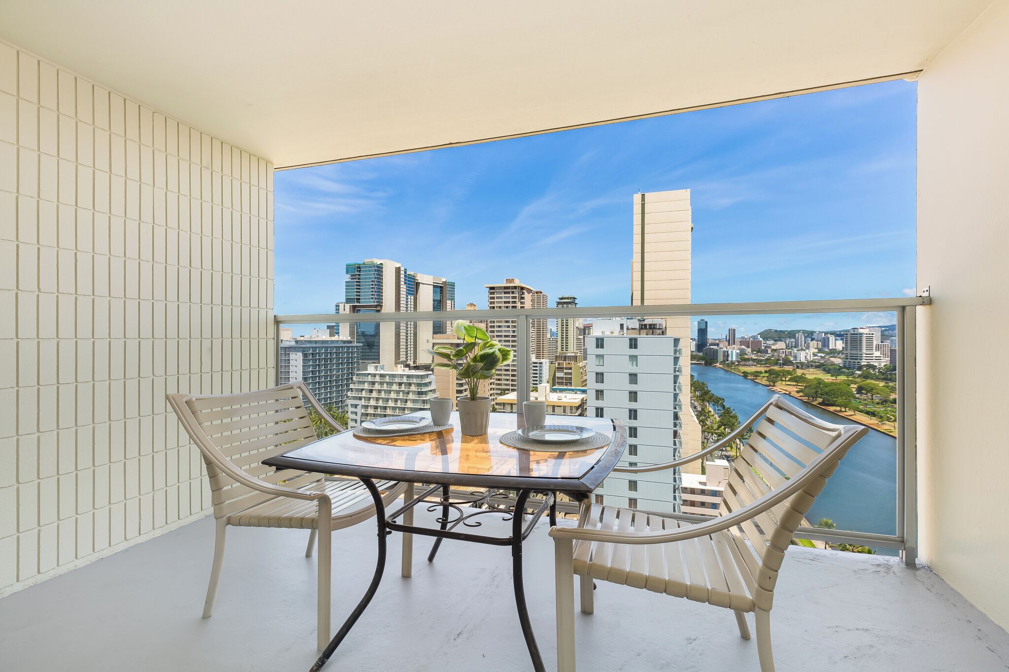 A balcony with a glass table and chairs, a potted plant, and a city view.