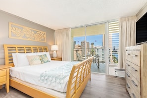 A hotel room with a bed, a nightstand, a lamp, a dresser, a balcony with a view of the city, and a large window. - Mountain & Golf Course Views Family Island Colony Condo & Lots of Amenities (Honolulu)