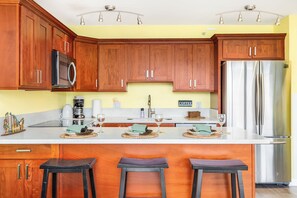 A kitchen with wooden cabinets, a stainless steel refrigerator, a coffee maker, and bar stools. - Golf Course-city View Condo at the Waikiki Banyan By the Beach & Restaurants (Honolulu)