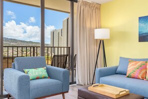 Un balcón con vista a un paisaje urbano y montañas, un sillón azul con un cojín floral, un sofá azul con un cojín estampado y una lámpara de pie.
