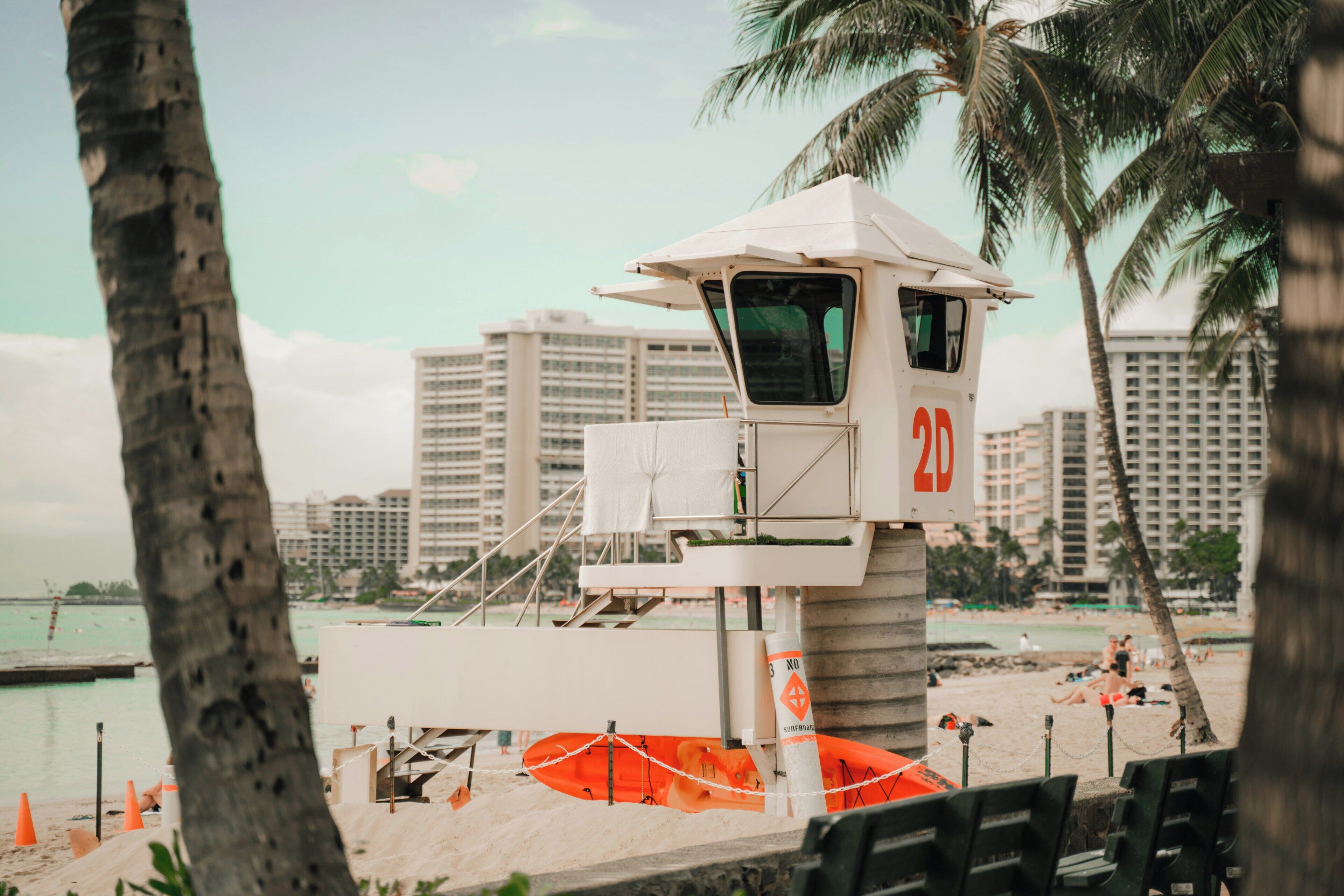 Une tour de surveillance, portant le numéro 20, surplombe une plage bordée de palmiers, avec une vue sur la ville en arrière-plan.