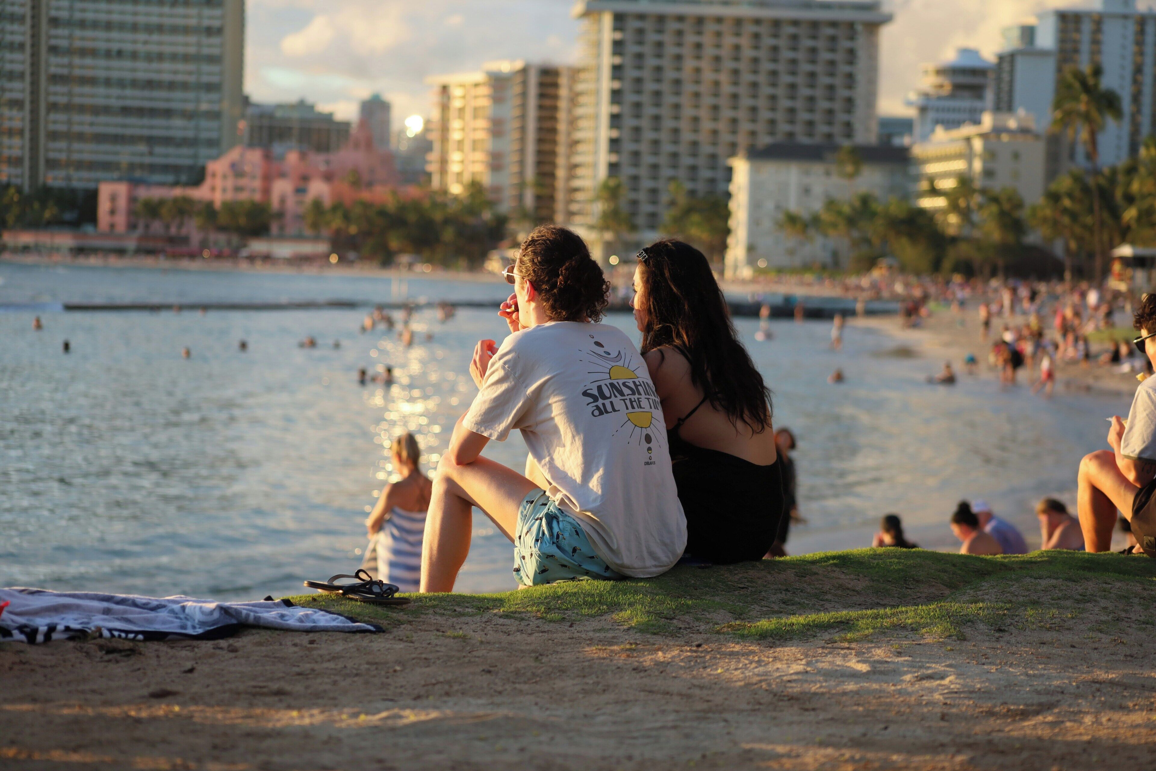 A beach scene with people sitting on the sand, a body of water in the background, and buildings in the distance.
