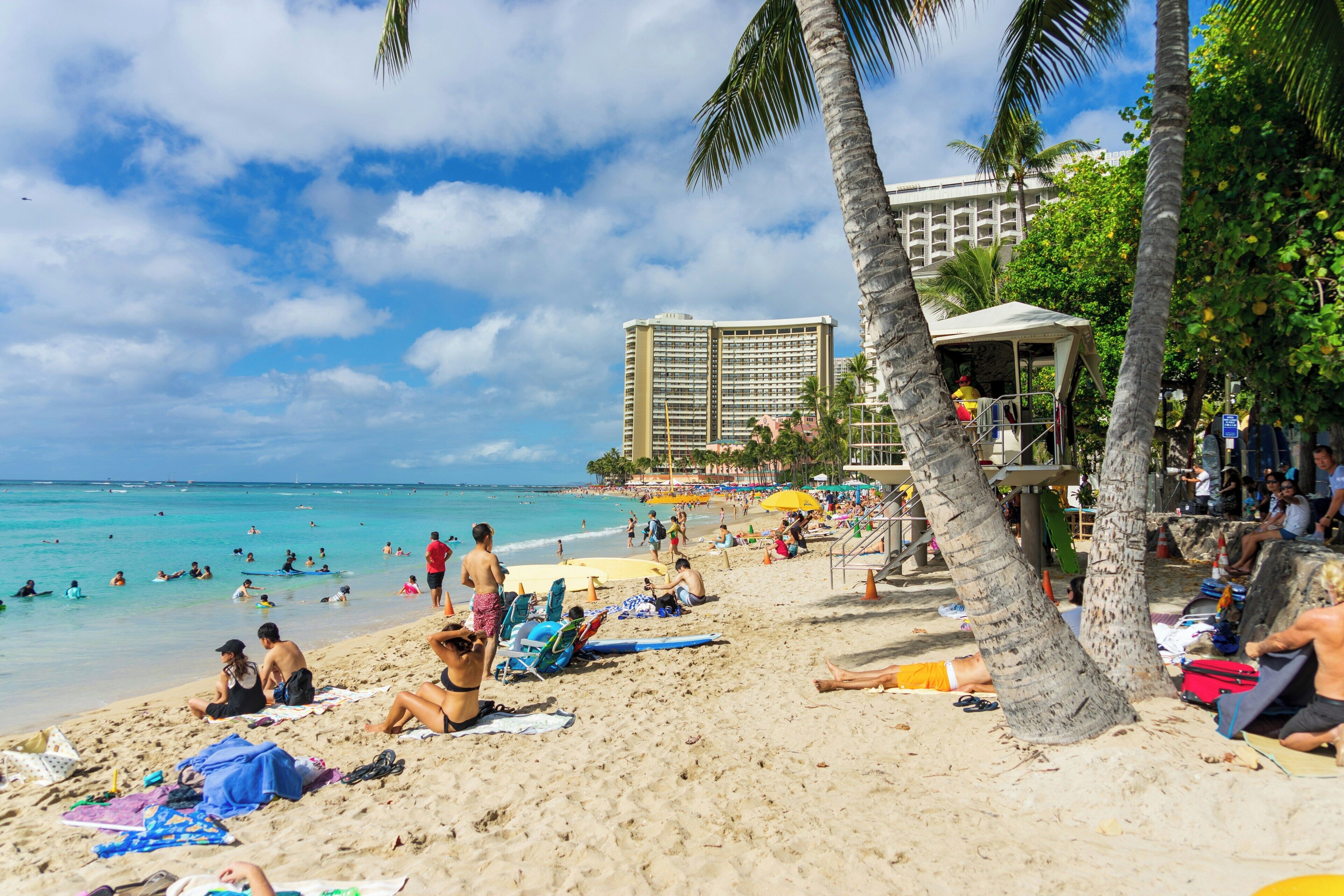 A beach with people sunbathing and swimming, a lifeguard tower, and a hotel in the background.