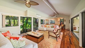 A spacious living room with a large sofa, a coffee table, and a dining area with chairs. There are large windows with a view of greenery outside.