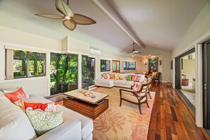A spacious living room with a large sofa, a coffee table, and a dining area with chairs. There are large windows with a view of greenery outside.