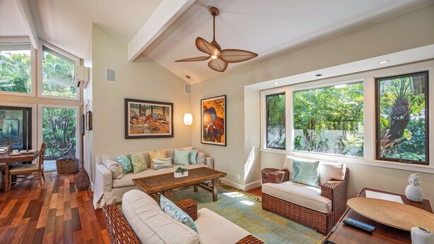 A spacious living room with a high ceiling fan, large windows with tropical views, a dining area, and a cozy seating arrangement.