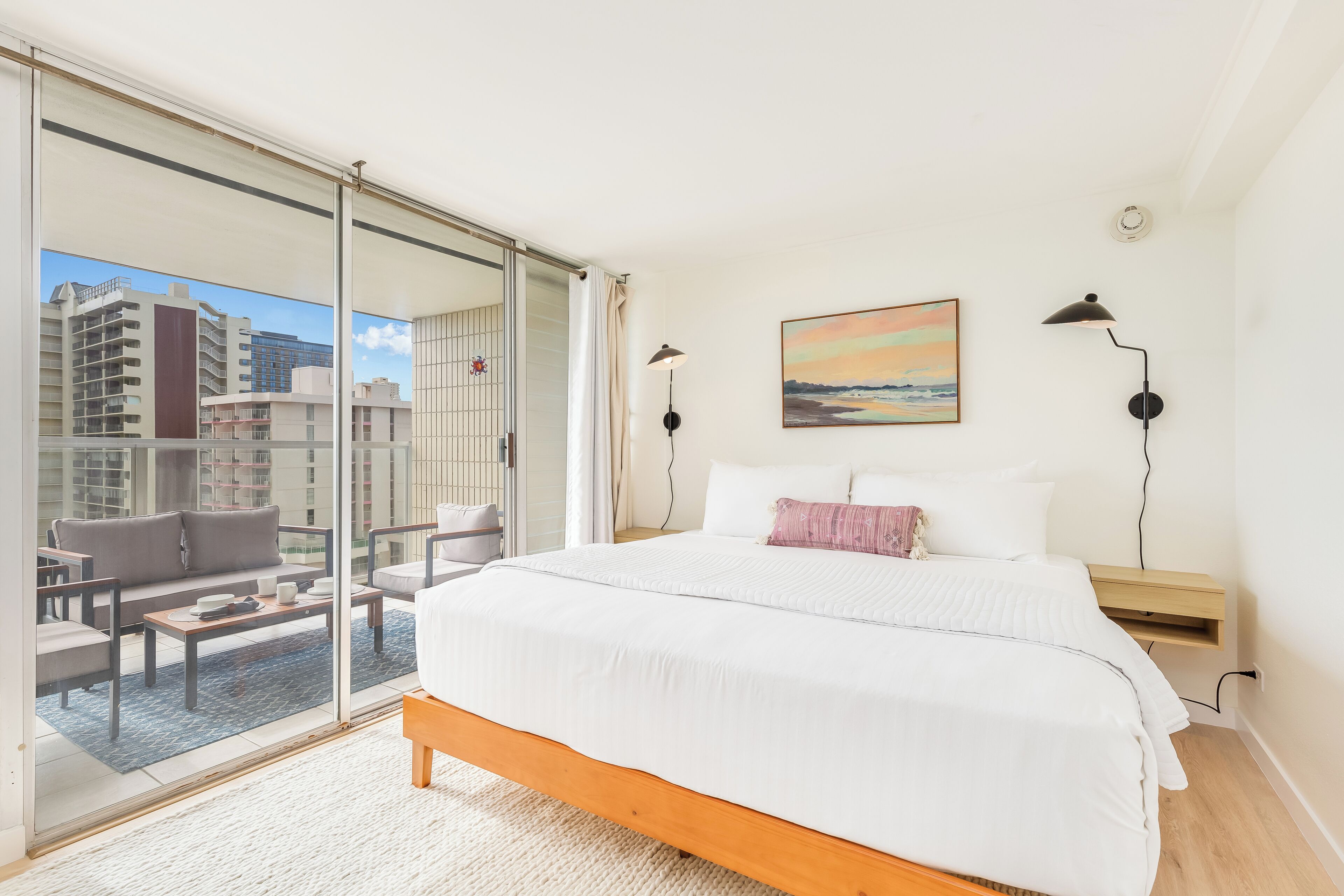 A hotel room with a large bed, a view of the city through floor-to-ceiling windows, a painting on the wall, and a balcony with seating.