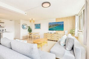 A modern living room with a sofa, a wooden coffee table, a TV, and a painting of a beach. - Golf Course & MT Renovated Condo at Island Colony & Spectacular Amenities (Honolulu)