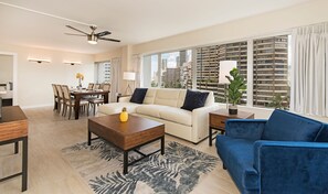 A modern living room with a sofa, coffee table, and blue armchair. There is a dining area with a table and chairs. The room has large windows with a city view. - Ilikai Hotel City View Condo Steps Away From Beach W/full Kitchen & Free Wifi (Honolulu)