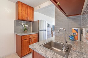 A kitchen with wooden cabinets, a stainless steel refrigerator, a granite countertop, a sink, and a potted orchid. - Ocean & Diamond Head Views Condo at Island Colony W/building Amenities (Honolulu)