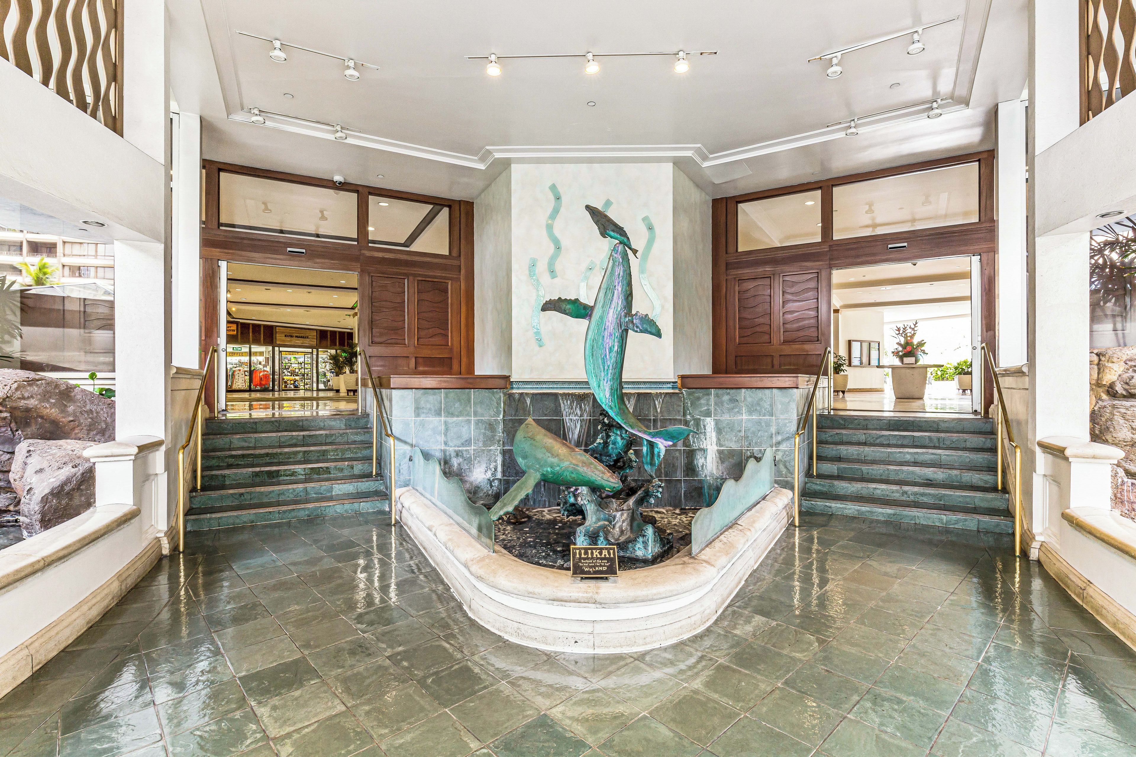 A lobby with a central fountain sculpture, a staircase, and a view of a commercial area.