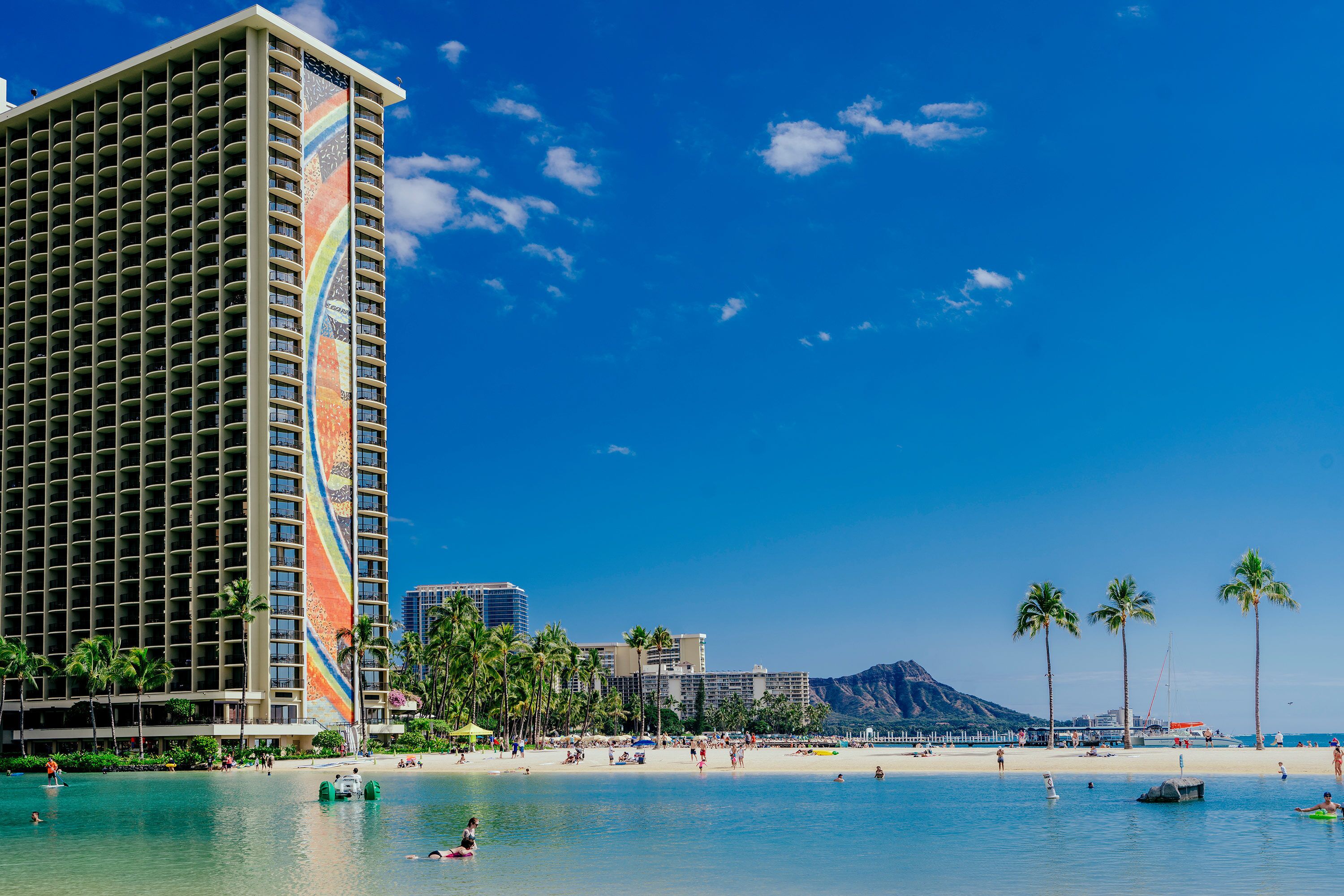 Un hotel frente a la playa con un edificio grande, palmeras y un cielo azul despejado.
