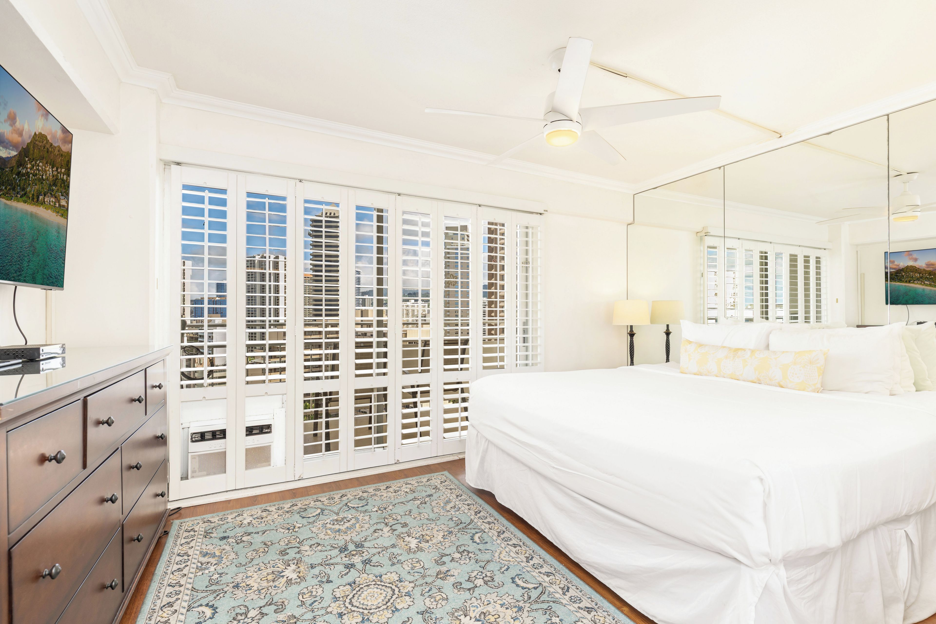 A bedroom with a bed, a dresser, a mirror, and a view of a beach through the window.