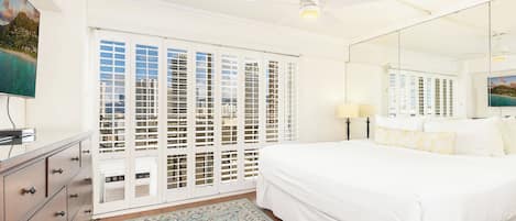 A bedroom with a bed, a dresser, a mirror, and a view of a beach through the window.