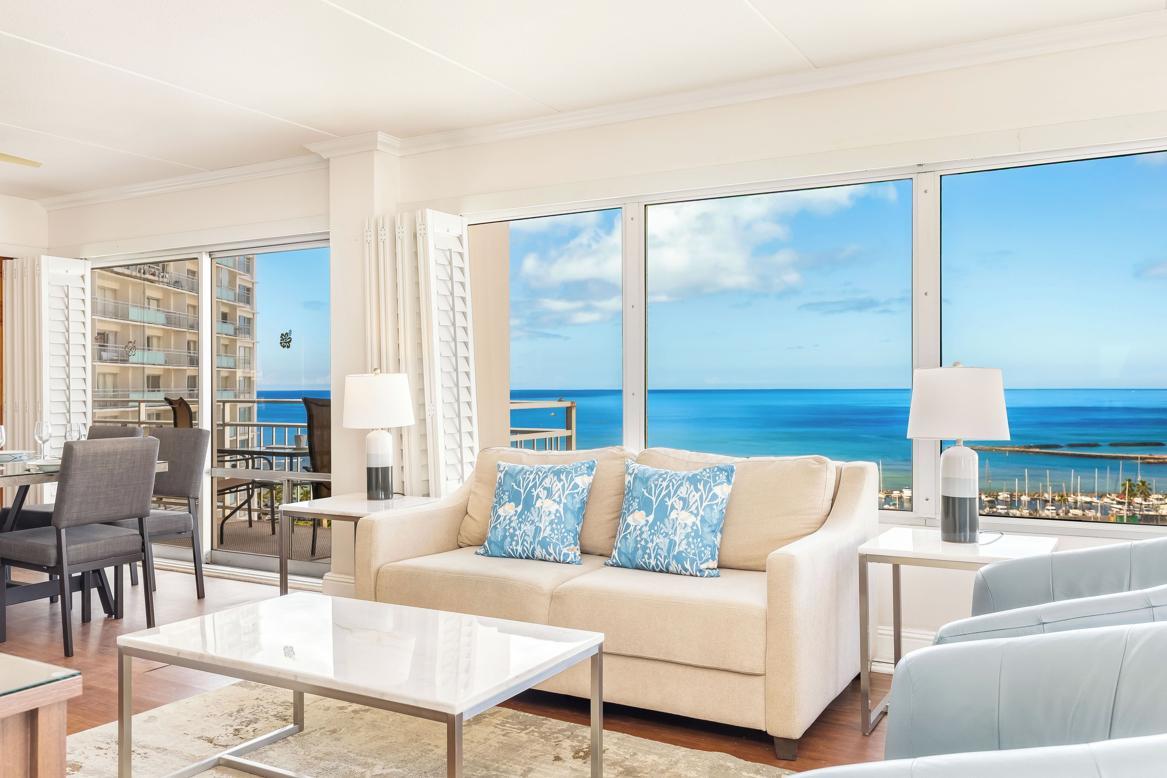 Ein modernes Wohnzimmer mit großem Fenster zum Meer, einer Couch mit gemusterten Kissen, einem Couchtisch und einem Essbereich mit Stühlen.