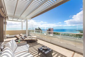 A spacious balcony with a view of the ocean and distant buildings, furnished with a sofa and a small table. - Luxurious Ocean View Panoramic PH at The Watermark W/spectacular Amenities (Honolulu)