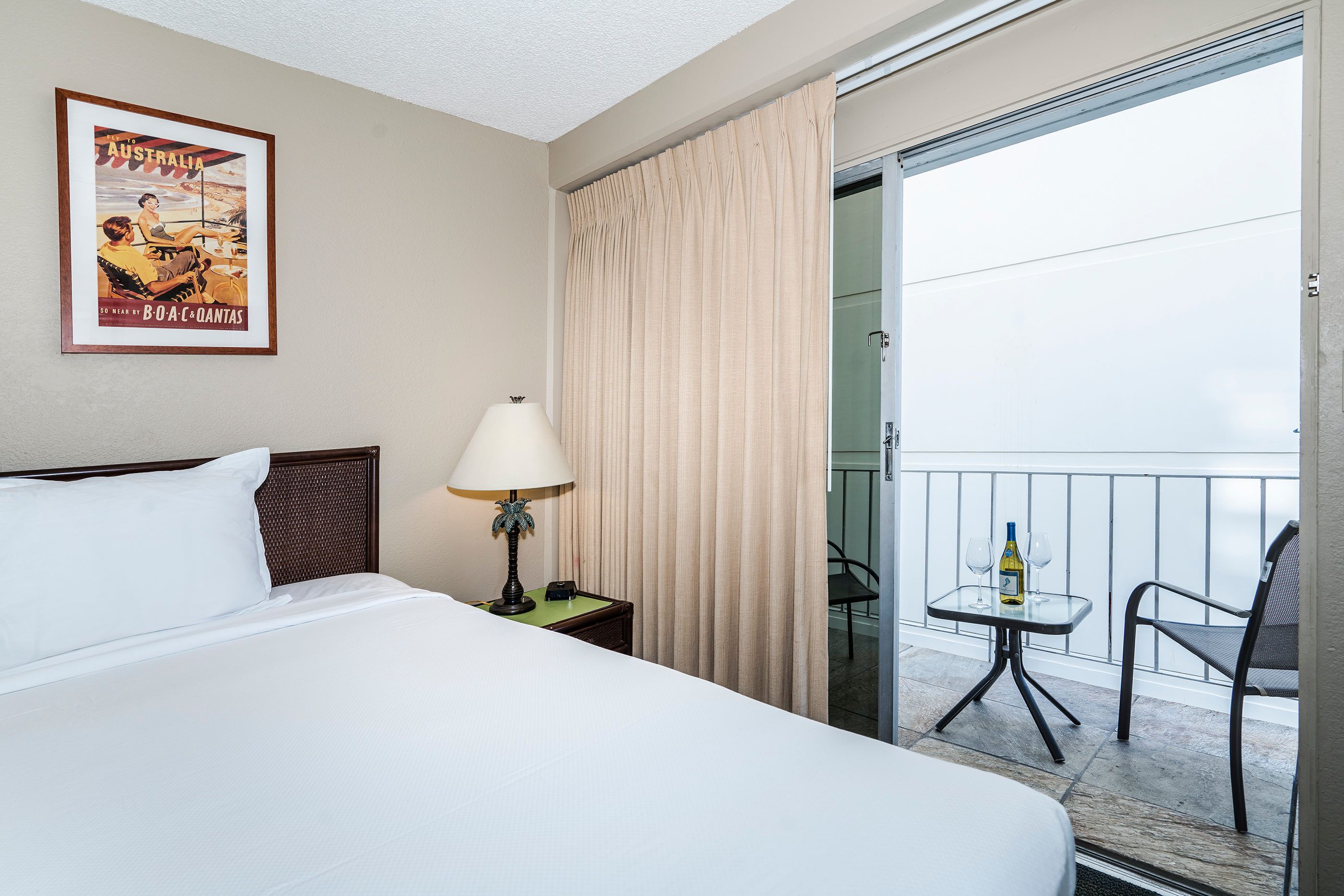 A hotel room with a bed, a nightstand with a lamp, a framed poster on the wall, a balcony with a table and chairs, and a sliding glass door.