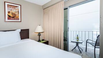 A hotel room with a bed, a nightstand with a lamp, a framed poster on the wall, a balcony with a table and chairs, and a sliding glass door.