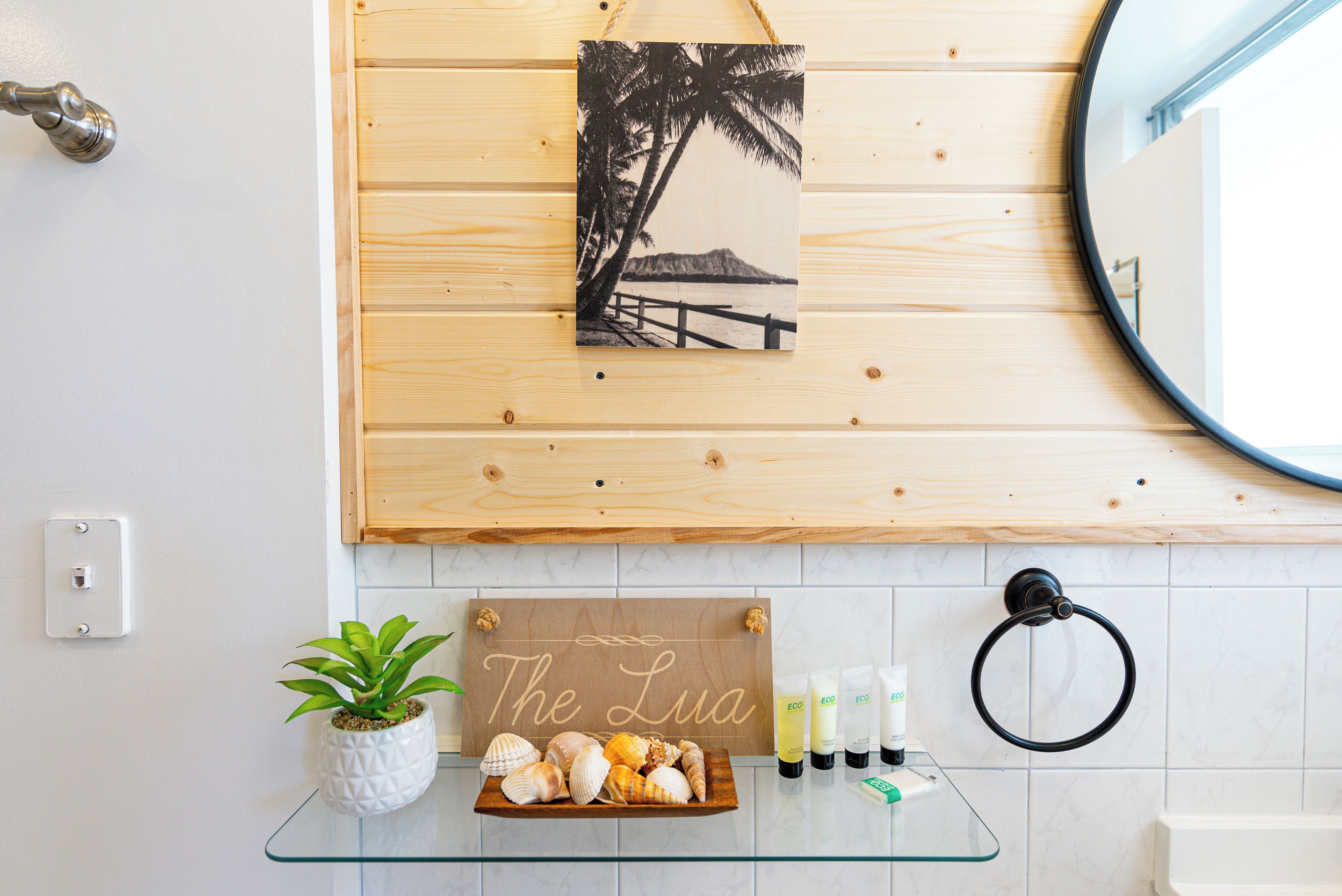 A bathroom with a wooden panel wall, a framed picture of a beach, a sign reading "The Lua", a glass shelf with a plant and seashells, and toiletries.