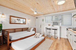 Una habitación con dos camas de madera, un ventilador de techo, una zona de cocina con un taburete de bar y un espejo grande en la pared.