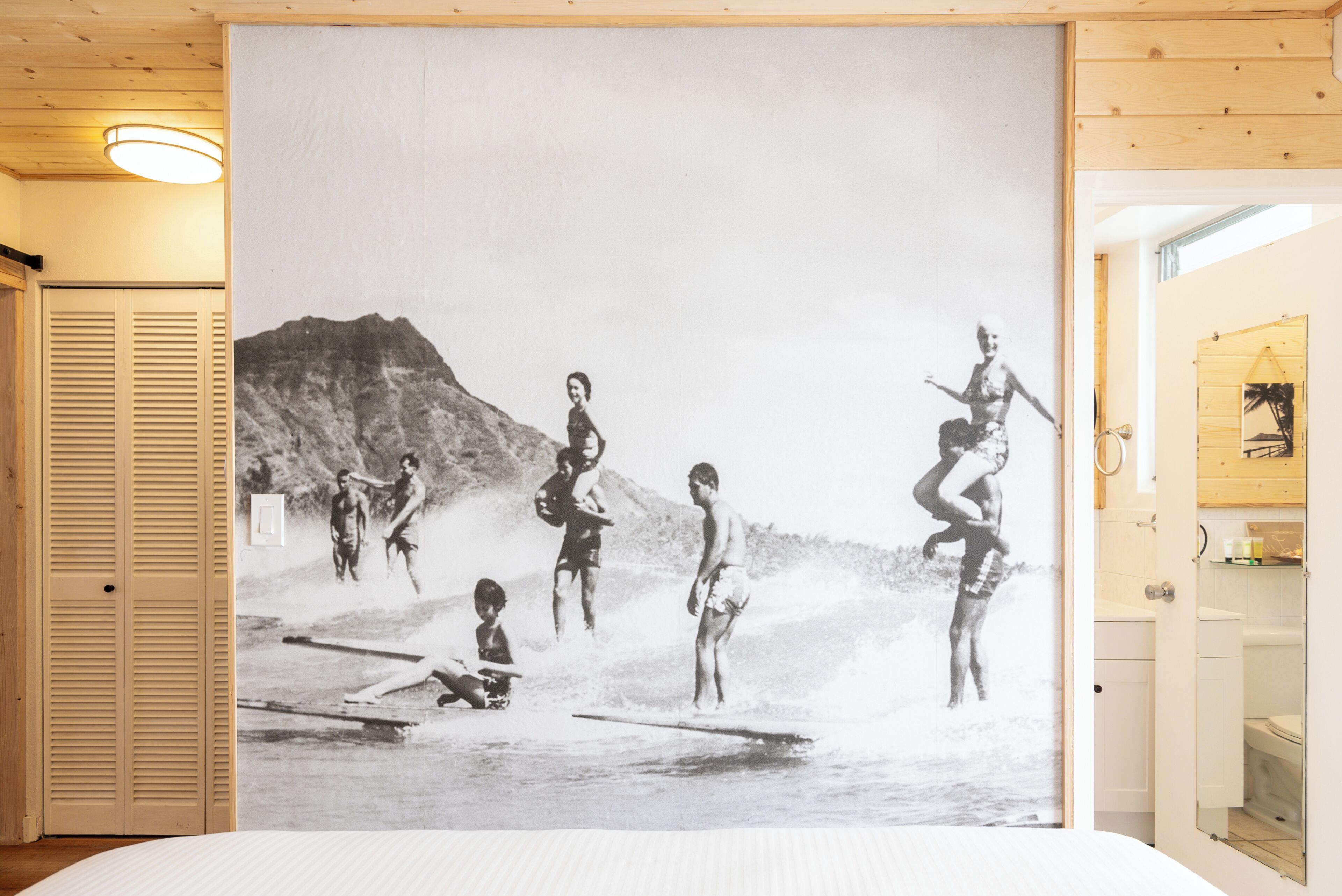 Una habitación con una fotografía grande en blanco y negro de gente surfeando, una puerta de madera y un baño a la vista a través de una puerta abierta.