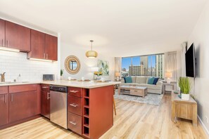 A modern kitchen with a dishwasher, a dining area with a table and chairs, a living room with a sofa and a TV, and a view of the cityscape through large windows. - Ocean & City Views Renovated Condo at Aloha Towers W/washer-dryer & Parking (Honolulu)
