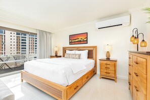 Ein Hotelzimmer mit einem großen Bett, einem hölzernen Schminktisch, einem Nachttisch mit Lampe, einer Wand-Klimaanlage und einem Balkon mit Blick auf die Stadt.