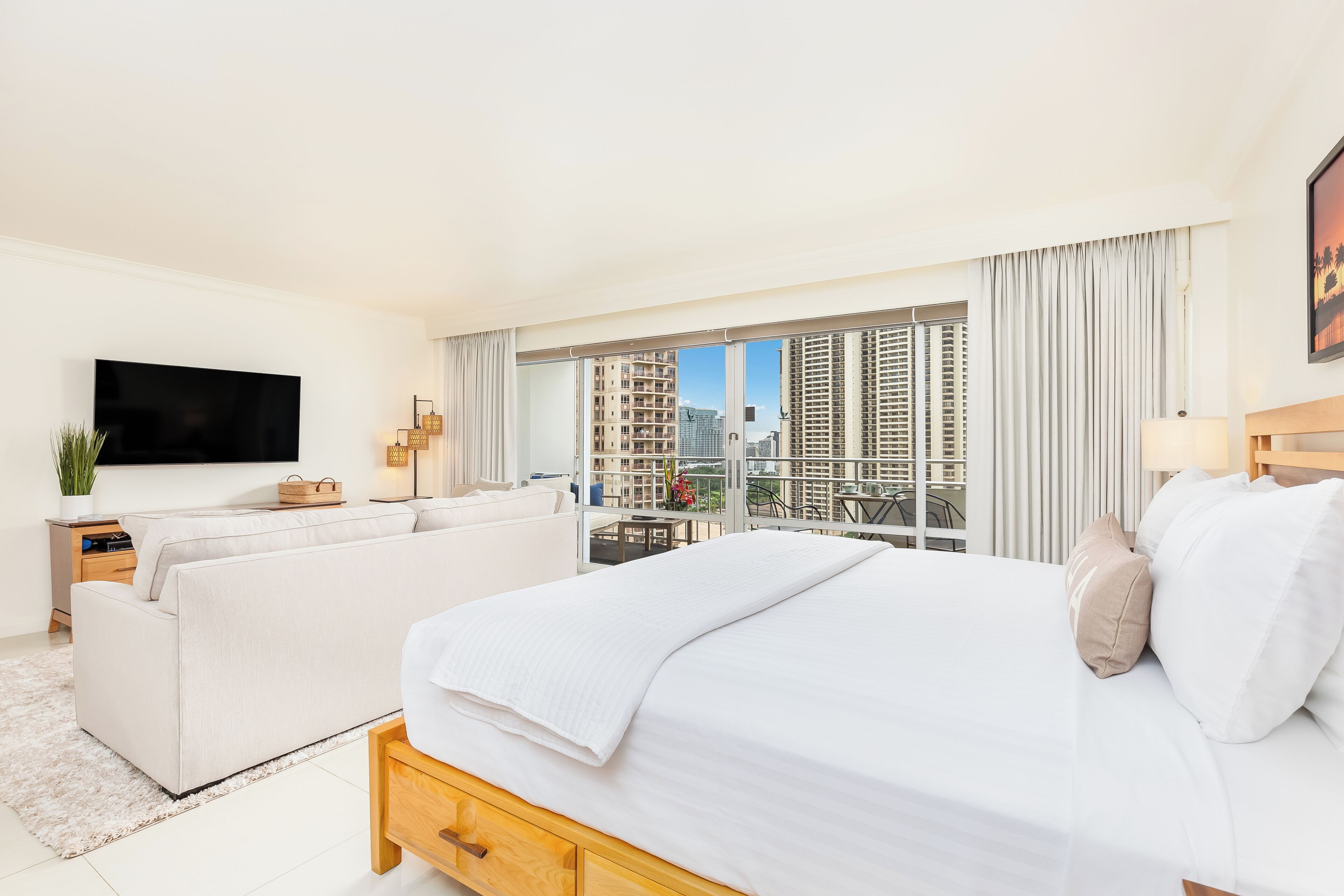 A hotel room with a bed, a sofa, a TV, a balcony with a city view, and a dining area.