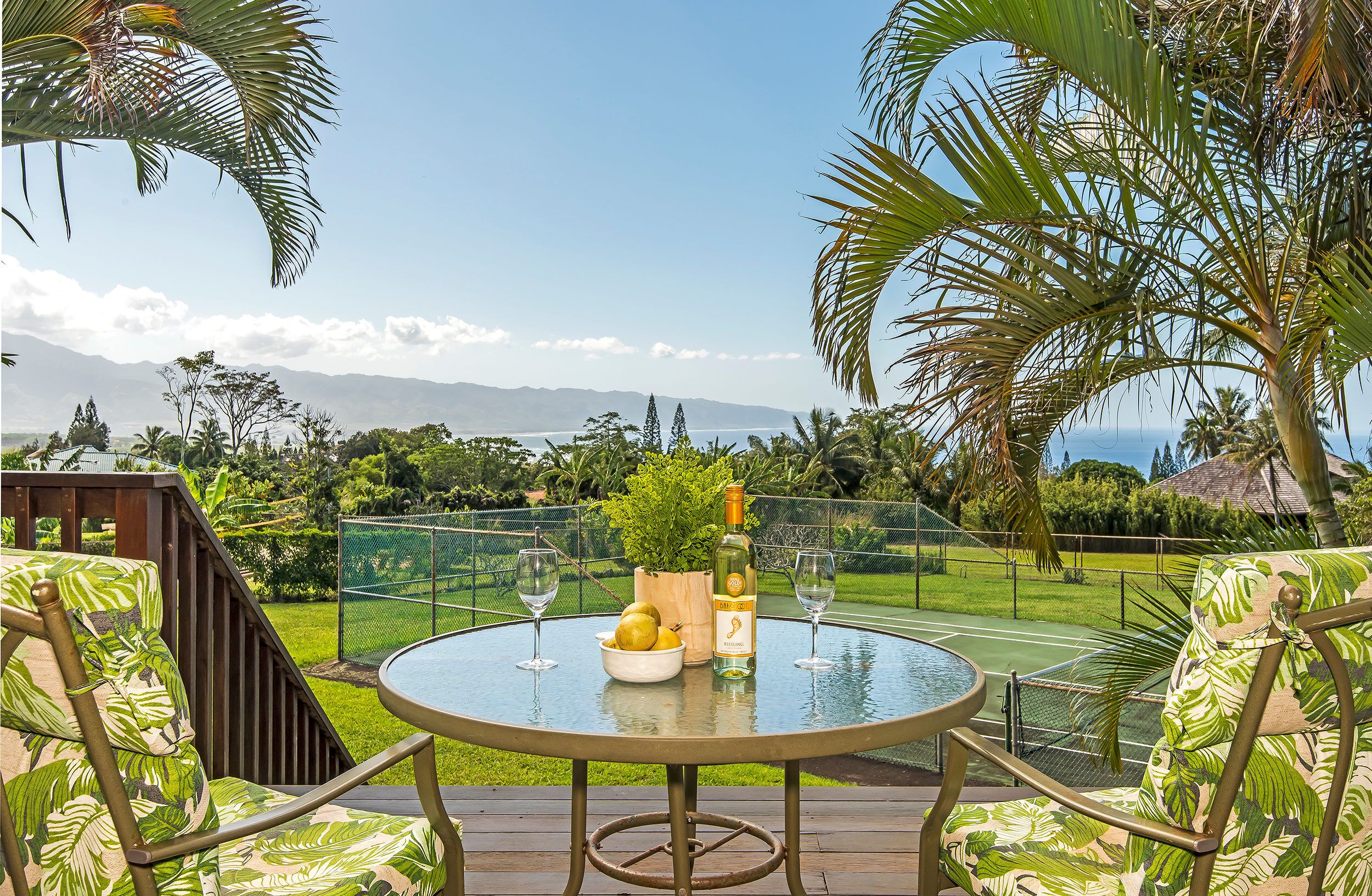 A round glass table with a bottle and glasses, surrounded by chairs with a tropical print, overlooking a lush garden and a distant mountain range.