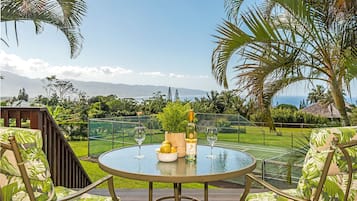 A round glass table with a bottle and glasses, surrounded by chairs with a tropical print, overlooking a lush garden and a distant mountain range.