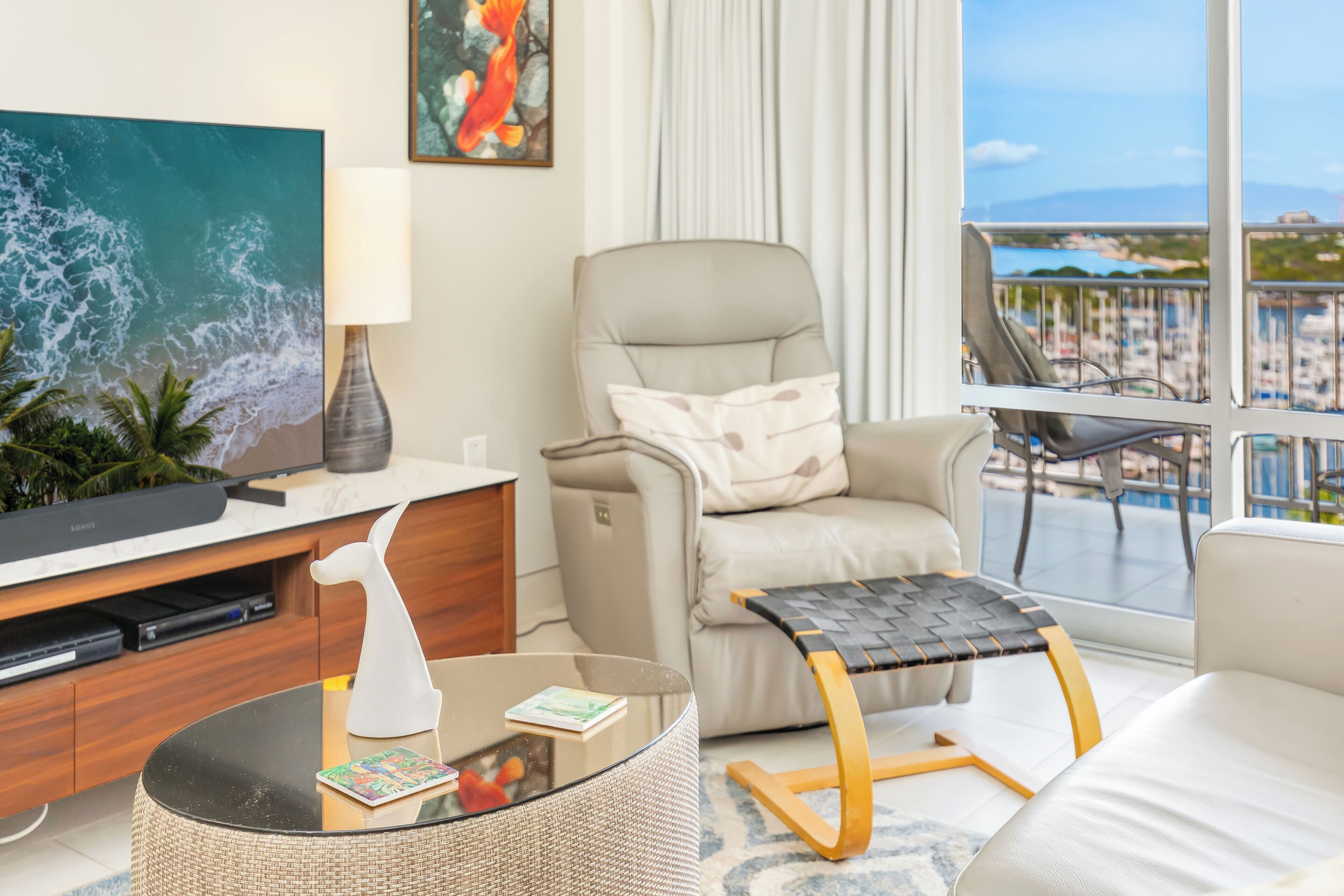 Un salón moderno con un televisor grande, asientos cómodos, una mesa de centro de cristal y un balcón con vistas al mar y a edificios.