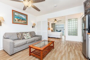 Un salon avec un canapé gris, une table basse en bois, un ventilateur de plafond, un tableau, et un coin cuisine avec un réfrigérateur.
