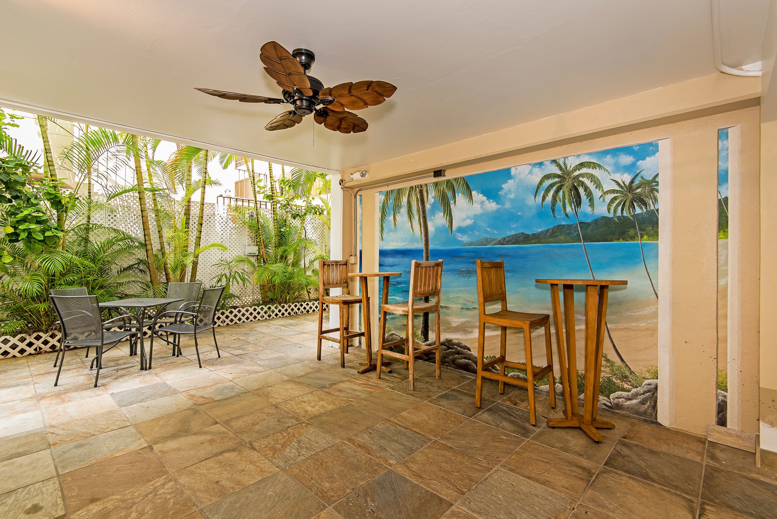 Um pátio externo com ventilador de teto, uma mesa com cadeiras e um mural com cena de praia.