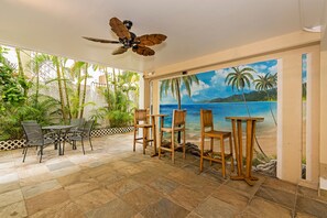 Um pátio externo com ventilador de teto, uma mesa com cadeiras e um mural com cena de praia.
