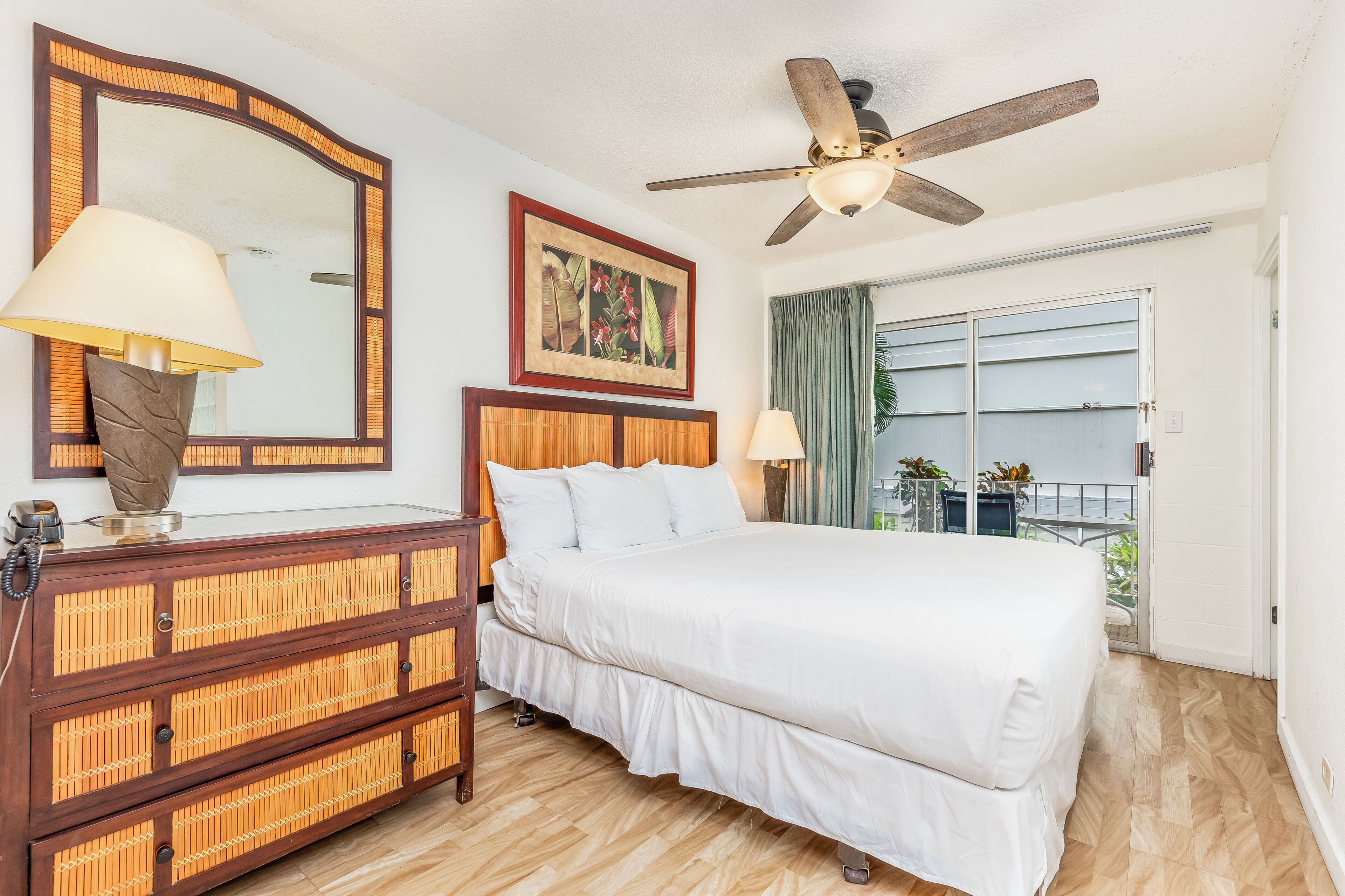 Une chambre d’hôtel avec un lit, une commode en bois, un ventilateur de plafond, un grand miroir et une fenêtre donnant sur un beau paysage.